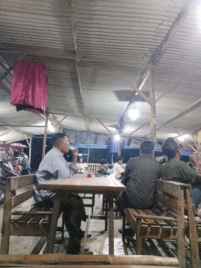 The image depicts an outdoor seating area of a casual eatery, likely a warung or similar establishment, in Sugihwaras, Indonesia. The scene is lit by artificial lights, suggesting it is evening or night. The structure has a corrugated metal roof supported by wooden beams and poles. A dark red fabric hangs from the ceiling on the left.
In the foreground, a man in a light blue shirt is seated at a wooden table, facing away from the camera and smoking. He appears to be in his 40s or 50s, with short, dark hair. His left elbow rests on the table, and he holds a cigarette to his mouth with his right hand. His attire is casual, with dark trousers.
Further back, several other individuals are visible. On the right side of the frame, two men are seated on a wooden bench, facing away from the camera. The man on the left wears a dark grey shirt, and the man on the right wears a greenish-brown shirt. They appear to be engaged in conversation or observing something.
In the middle ground, a person in a striped shirt is standing near a counter or cart, possibly preparing food or serving customers. A motorcycle is parked to the left of this person. Other patrons are seated at tables in the background, some appearing to be younger individuals. The ground appears to be concrete or a similar hard surface, with some puddles or wet patches visible, indicating recent rain or cleaning. The overall atmosphere suggests a relaxed, informal gathering place for local residents.
