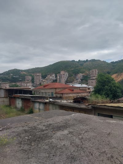 The image shows a view of apartment buildings built on a steep, green mountainside. The buildings are of varying heights and architectural styles, with some appearing older and more worn than others. The mountainside is covered in lush vegetation, with trees and bushes growing around the buildings. The sky is overcast and appears to be a dull, gray color.

In the foreground, there is a concrete structure with a rough texture, possibly a road or a wall. To the left of the concrete structure, there are several garage-like structures with metal doors. Some of these doors are rusted, and vegetation is growing around them. A red-tiled roof is visible on one of the buildings, adding a touch of color to the otherwise muted palette of the scene.

The overall impression is one of a somewhat weathered urban environment nestled into a natural landscape. The activity taking place is not evident, but the scene suggests a residential area. The time of day is difficult to ascertain due to the overcast sky, but it appears to be daytime. There are no people visible in the image. Some faint numbers appear on one of the garage doors. The location context of Kapan, Armenia, suggests a mountainous region, which is consistent with the visual elements.