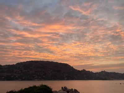 The image depicts a landscape at Hale Bandi Harlapura, India, during either sunrise or sunset. The sky is largely covered by a dense formation of altocumulus clouds, presenting a dominant spectrum of orange and pink hues, intermingled with lighter grey and pale blue towards the top. A dark, rugged mountain range or series of rocky hills, heavily dotted with boulders and sparse vegetation, forms the horizon line, silhouetted against the illuminated sky. Below the hills, a calm body of water occupies the mid-ground, reflecting the warm, reddish-orange light from the sky. In the immediate foreground, a cluster of dark green bushes is visible on the left, and several large, smooth, grey boulders are situated at the water's edge on the right. No human figures, animals, or man-made structures are discernible within the scene.