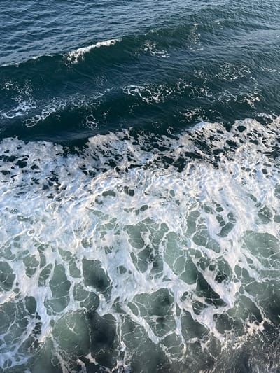 The scene depicts a dynamic expanse of open seawater in the vicinity of Kato Karioutes, Greece, viewed from an elevated perspective. The upper third of the image features dark teal to deep blue water with visible surface undulations and a minor wave crest. A distinct horizontal boundary, approximately mid-frame, separates this calmer water from an extensive area of white seafoam and turbulent water. This foamy region, occupying the lower two-thirds of the image, displays complex, irregular patterns of aerated white water and lighter blue-green water. The foam is consistent with breaking waves or the wake of a vessel. No individuals, vessels, or man-made objects are present within the frame. The environment is entirely marine, characterized by active water movement and foam generation.