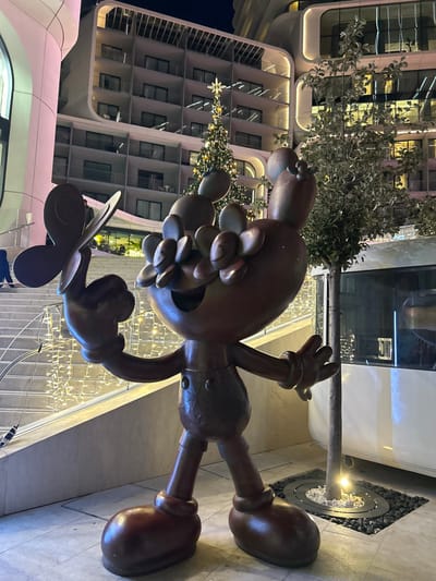 The scene depicts an outdoor nighttime view in Saint Julian's, Malta, featuring a bronze-colored statue against a backdrop of modern architecture and festive lighting.
In the foreground, a large, stylized, bronze-colored statue of a cartoon-like character stands centrally. The figure possesses a large head adorned with rounded, petal-like elements, a wide smiling mouth, and large, gloved hands. Its left hand is open with the palm forward, while its right index finger is raised, upon which a stylized butterfly with two distinct wings rests. The statue wears patterned overalls with two star-shaped depressions on the torso.
To the left and behind the statue, a descending staircase is visible, illuminated by numerous vertical strings of small, warm-toned lights. To the right of the statue, a slender tree with sparse foliage grows from a circular ground planter filled with a ring of black stones surrounding an inner circle of white stones, lit by an upward-facing ground light. A reflective surface, possibly a large window, is partially visible further right.
The background is dominated by a multi-story, contemporary building with a light-colored, possibly white, facade featuring rounded architectural elements and numerous balconies with glass railings. The building's interior is illuminated, casting a warm glow from its windows. A tall, conical Christmas tree, decorated with numerous golden lights and topped with a large star, stands in front of the building. The sky above is dark, indicating night.