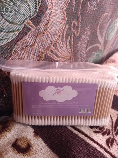 This is a close-up, eye-level shot of a package of cotton swabs with wooden sticks. The package is clear plastic, revealing the white cotton swabs neatly arranged inside. A purple label is affixed to the front of the package, featuring a cloud graphic with a stylized figure wearing a hat. Text in Russian is visible on the label, indicating the product is "Cotton swabs on a wooden base, 500 pcs." A barcode is also present on the label. The background consists of a patterned textile, likely a blanket or upholstery, with a soft, woven texture in muted colors. The image is brightly lit, with no visible shadows, suggesting an indoor setting. There are no people or discernible landmarks to indicate a specific location within Monastirische, Russia.