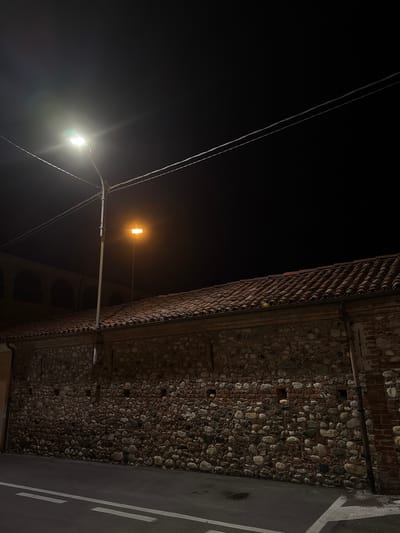 The image depicts a night scene on a street in Lagnasco, Italy. A tall street lamp illuminates a section of a rough stone wall with a tiled roof. The wall is constructed of irregular stones and some brickwork, with small openings or recesses. The street in the foreground is asphalt with white lane markings. In the background, the upper portion of a building with arched windows is visible, suggesting it might be an older structure. The sky is dark, and power lines are visible stretching across the frame. The overall impression is one of a quiet, dimly lit street at night.