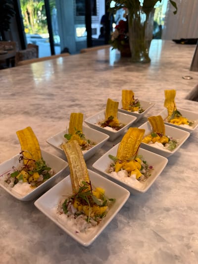 Nine individual appetizers are meticulously arranged in three rows on a light-colored countertop with grey veining, indicative of marble. Each appetizer is presented in a small, square, white ceramic bowl. The contents of each bowl consist of a white creamy base, diced light-colored material, diced yellow fruit (consistent with mango), green microgreens, and a sprinkling of reddish-brown spice. A single, upright, rectangular fried plantain chip, yellow-orange in color, garnishes each portion. In the background, a tall, clear glass vase holds green foliage and several red flowers. Beyond the counter, blurred reflections and outlines of glass doors or large windows reveal outdoor foliage, suggesting an open and well-lit interior. No individuals are visible. The scene depicts a prepared food display in what appears to be a modern, upscale residential, culinary, or hospitality setting. The specific location within Naples, Florida, United States of America, is not identifiable from the visual information provided.