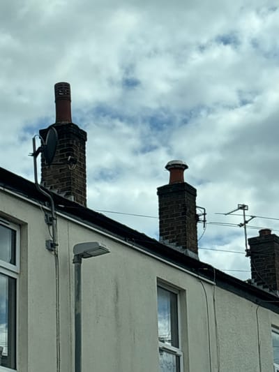 The image presents an upward view of a building exterior, likely a house, against a backdrop of a cloudy sky. The building is a light-colored structure with multiple windows. Visible on the roof are three brick chimneys, each with a distinctive top. An antenna is fixed on one of the chimneys. A security light fixture and some exterior cabling are present on the building's wall. The location is suspected to be in the city of South Ribble, United Kingdom. No people or interactions are visible in the frame.