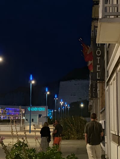 The image depicts a nighttime scene in Saint-Valery-en-Caux, France, with a view of the waterfront and a prominent building labeled "CASINO" in neon blue. In the foreground, several people are standing on a paved area. Two individuals are facing away from the camera, one wearing a black top and a patterned skirt, and the other wearing a dark green short-sleeved shirt and light-colored pants. Another person, partially visible, is wearing orange shorts and a dark top. Behind them, several tall lamp posts with blue lights line the walkway. Further in the background, the Casino building is visible with illuminated windows and signage. To the right of the image, a building with a sign that reads "HOTEL" in vertical lettering is prominent, with flags displayed above it. The overall environment appears to be a public promenade or street.