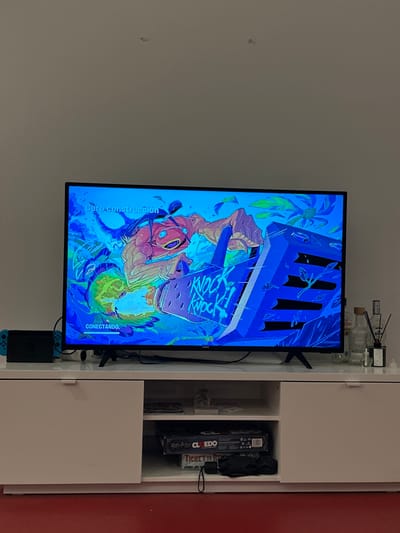 A television displays artwork featuring vibrant blues and a figure, seemingly from the game "Knock Knock." A Nintendo Switch dock is connected to the bottom of the screen. A white shelf unit sits beside the TV. On top of the shelf are various clear glass bottles. Inside the shelf is a Clue board game and other miscellaneous items. The image is oriented vertically.