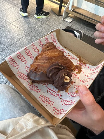 A chocolate croissant sits in a brown cardboard box lined with white paper printed with "Bloom Blood" in red. The croissant is topped with a swirl of chocolate cream and a hazelnut. A person's hands hold the box, with parts of their black clothing visible. The floor is tiled in light gray and white. Another person's legs are seen in the background, wearing black pants and New Balance sneakers.
