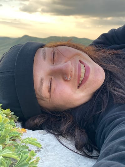 A close-up, low-angle, upside-down shot shows a young woman with her eyes closed, smiling broadly, lying on a rock with green plants in the foreground. She is wearing a black beanie and a black hooded sweatshirt. Her reddish-brown hair is spread out around her. A silver hoop nose ring is visible. In the background, rolling hills are visible under a cloudy sky with soft, warm light. The location is stated as Абзаново, Russia.