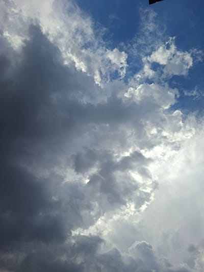 The image captures a dynamic sky filled with a mix of dark, ominous storm clouds and brighter, scattered cumulus clouds against a backdrop of clear blue sky. The lighting suggests a transition from bright sunlight to approaching darkness, with some areas of the clouds catching the sun's rays, creating a dramatic contrast. The scene is devoid of any identifiable structures or people. Based on the provided context, this sky was observed in Dolna Malina, Bulgaria.