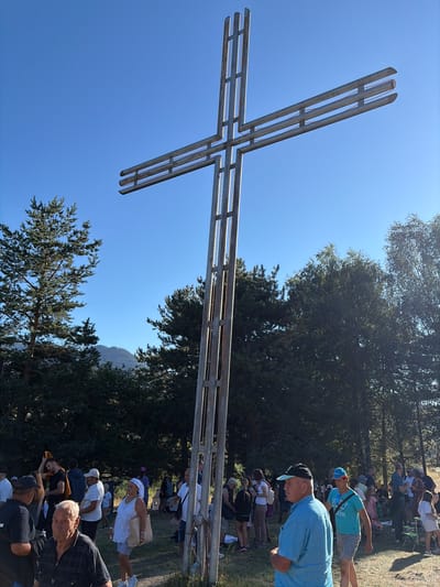A large, metallic cross stands prominently in a natural outdoor setting, against a clear blue sky. The cross is constructed from slender metal beams, forming a cross shape with a vertical beam extending upwards and a horizontal beam extending outwards. In the background, a crowd of people is gathered, suggesting a public event or gathering. The environment appears to be a wooded area, with pine trees visible in the midground and a mountainous landscape faintly discernible in the distance. The lighting indicates it is daytime, likely late morning or early afternoon, with the sun casting shadows. The people are casually dressed, some appear to be conversing, while others are looking towards the cross or engaging with the event. The overall scene suggests a religious or commemorative occasion taking place in a scenic, possibly mountainous, location such as the vicinity of Shiroka Laka, Bulgaria.