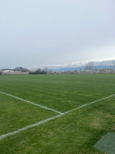 The image depicts a grassy soccer field in Voysil, Bulgaria. White lines mark the field boundaries. In the background, there are trees, a building, and a hazy sky. Beyond the trees is a mountain range.