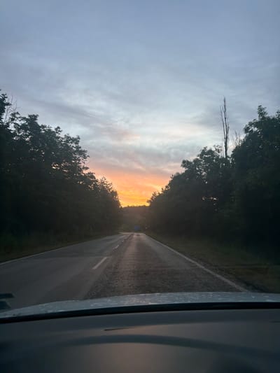 This is a first-person perspective, wide-angle photograph taken from inside a vehicle, looking forward. The scene captures a road with trees lining both sides, leading towards a sunrise. The sky is a gradient of pale blue to grey, with streaks of orange and pink from the rising sun visible in the distance. The road appears wet, possibly from recent rain, and has a white line marking its center. The foreground shows the dashboard and windshield of the vehicle. The environment suggests a rural or semi-rural setting, possibly on the outskirts of the city of Хаджийско, Bulgaria. There are no people or animals visible in the frame. The overall mood is serene and contemplative, indicative of an early morning drive.