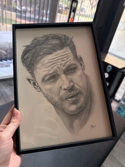 A framed pencil sketch of a man's face is held up by a person's hand. The sketch is tilted to the side within the black frame. The man in the sketch has furrowed brows, closed eyes, and a beard. His hair is dark and textured. A signature, "Mancoo" followed by "2025", is visible in the upper right corner of the sketch. The background of the image shows what appears to be a room with blinds on a window to the left, suggesting an indoor setting during the daytime. The overall mood of the sketch is intense and contemplative.
