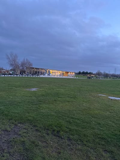 The image depicts an outdoor scene at dusk or early evening within The Helix Park, Falkirk, United Kingdom. The foreground consists of a large expanse of green grass, showing visible patches of mud and several small puddles.
In the midground, a modern, single-story building with a predominantly glass facade is prominently featured. The word "CAFE" is legible in large, white lettering on the building's glass front, which is brightly illuminated from within by warm yellow lights. Inside the cafe, several indistinct human figures are visible. To the left of the building, a cluster of bare-branched trees, consistent with birches, stands. Immediately in front of and to the left of the cafe, a series of dark picnic tables with benches are arranged on a paved surface. A large, pale-colored sculpture, identifiable as the head of one of The Kelpies, is positioned directly in front of the cafe's main entrance.
To the right of the building, a paved pathway is visible, along which several human figures are walking. A string of decorative outdoor lights is suspended above this pathway. In the far background, distant trees and at least two large metal electricity pylons are discernible against the horizon. The sky is heavily overcast with a uniform grey-blue tone, indicating an absence of direct sunlight.