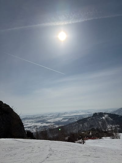 The image depicts a panoramic winter landscape from a snow-covered mountain slope in Zelenaya Polyana, Russia. The sky, occupying the upper two-thirds of the frame, is light blue with a brilliant, circular sun positioned in the upper center, generating a prominent lens flare and illuminating the scene directly. A linear contrail from an aircraft extends diagonally across the sky from the upper left to the lower-mid left. The foreground features a wide expanse of disturbed snow, indicating a ski slope with multiple ski tracks. To the far left, a large, dark rock formation rises, partially framing the view. The midground showcases snow-dusted mountains and hills sparsely covered with bare deciduous trees and some evergreen foliage. A cluster of small, red-roofed buildings is discernible on a distant slope to the right. A distinct green lens flare artifact is present in the lower-midground. The background comprises an expansive, hazy vista of snow-covered plains and rolling terrain stretching to the horizon under a pale sky. No individuals are visible.