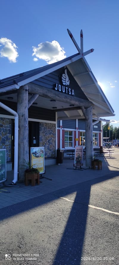 The image shows the exterior of a building in Joutsa, Finland, with the name "JOUTSA" prominently displayed above the entrance. The building has a rustic architectural style, featuring natural wood pillars and stone cladding on the lower portion of the facade. The roof is dark-colored with white trim, and there are decorative wooden elements at the peak. A large sign with a thumbs-up graphic is part of the building's branding. Several other signs are visible on the sidewalk in front of the building, one of which advertises a hotel. Picnic tables and trash receptacles are also present. The ground is paved with interlocking bricks, and a portion of the foreground is asphalt. The scene is illuminated by sunlight, casting shadows across the pavement. The sky is blue with scattered white clouds, and some trees are visible in the background. The date and time stamp "2026/6/30 20:04" is visible in the lower right corner of the image, along with camera information.