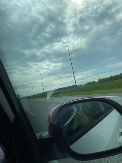 The image shows a view from inside a car, looking out the side window and into the side mirror. The sky is overcast with grey clouds. Outside the car, a highway stretches into the distance, lined with green fields and trees on either side. Several tall streetlights are visible along the highway. In the side mirror, a reflection of a person holding a phone is visible, seemingly taking the photograph. The person appears to be a woman, with dark hair, and is wearing a dark top. She is looking at the phone in her hand. The interior of the car, including part of the steering wheel and the dashboard, is also visible in the reflection. The scene appears to be a travel scene on a highway, possibly in a rural area given the presence of fields and trees. The specific location in the city of Pervomaiskoe, Russia, cannot be determined from the visual information provided in the image.