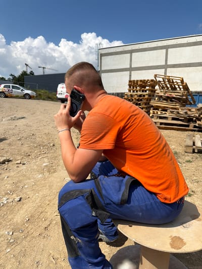 A person wearing a bright orange t-shirt and blue work overalls is crouching down on a dirt ground. They are holding a smartphone in their right hand, appearing to be taking a photo or video. The person has short, light brown hair. The scene is outdoors, with a clear blue sky and scattered white clouds visible. In the background, there is a large, light-colored building with a grid-like structure. Piles of wooden pallets are stacked near the building. To the right of the person, a large, light-colored cylindrical object, possibly a spool of cable, is partially visible. Further in the background, there are several white vans with some text, and what appears to be construction cranes. A white car with colorful branding is also visible in the foreground. The lighting suggests it is daytime, likely sunny. The overall setting appears to be a construction site or industrial area. The person's posture and focus on the phone suggest they are engaged in documenting something on the site.