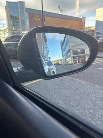 The image is a close-up, low-angle, exterior shot of a car's side mirror, reflecting a street scene. The mirror housing is black plastic and appears slightly dirty. The reflection shows a street with buildings on either side, a white car parked in the middle ground, and a grey car in the background. One building in the reflection has the word "CINEMA" visible in gold lettering. The street surface is asphalt with yellow painted lines. The sky is partly cloudy. The image is taken from inside a car, with the interior of the car visible in the foreground.
Based on the visible signage ("The Reel Cinema" and the architecture), the location is the area around the entrance to The Reel Cinema in Cork, Ireland, which is situated in the city center, likely on a street such as Great George's Street or a nearby road with similar urban development. The specific reflection in the mirror likely captures a street adjacent to the cinema, given the perspective.