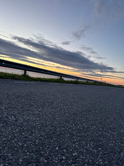 The image depicts a road with a guardrail running alongside it. Beyond the guardrail, a body of water is visible, with a sunset occurring in the background. The sky is filled with clouds, tinged with yellow and orange hues from the setting sun. The road surface is asphalt, with white lines marking lanes. The grass on the side of the road appears green and somewhat overgrown. The scene is quiet and serene, suggesting a location at the edge of a city or town. It's possible this is in Paldiski, Estonia, a port city known for its coastal scenery and historic sites.