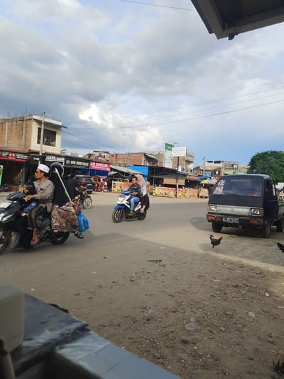 In Gampongpaya, Indonesia, on a dusty, outdoor street, a black, two-door pickup truck with a license plate reading "BL 8178 ZD" is parked facing left. A black and white rooster stands in front of the truck. In the middle distance, two people ride a blue and white motorcycle, the driver wearing a blue shirt and jeans, and a passenger wearing a white hijab and blue shirt. To the left, three people, two adults and a child, ride a black motorcycle. The adult driver wears a white cap and a patterned shirt, while the adult passenger wears a black headscarf and a patterned skirt. The child is seated in front of the adult driver. Further left, a building with a red brick facade and exposed concrete supports is visible, with various shops and signs lining the street. The sky above is cloudy, with patches of blue.