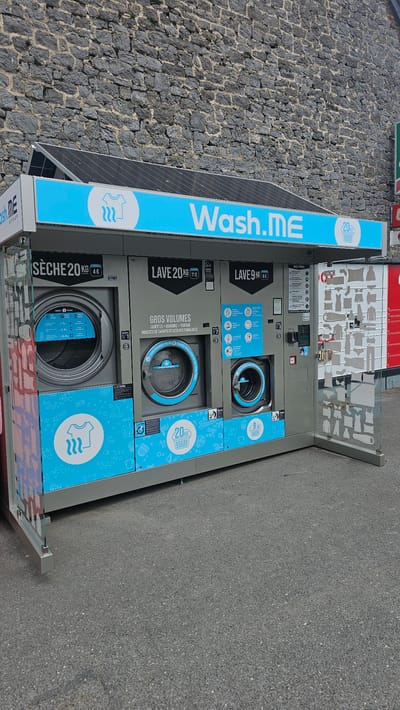 The image shows a row of self-service laundry machines at a "Wash.ME" facility. The machines are located outdoors, against a stone wall. The facility appears to be situated in the town of Boussu-lez-Walcourt, Belgium.
The machines are labeled with their capacities in kilograms and prices. There is a dryer labeled "SÈCHE 20KG 4€", a washing machine labeled "LAVE 20KG 11€" which is described as suitable for "GROS VOLUMES" (large volumes) like duvets, pillows, and family linens, and another washing machine labeled "LAVE 9KG 6€". The machines are primarily gray with blue accents and graphics, including images of clothes and laundry baskets. Some of the machines have digital displays.
To the right of the washing machines, there is a glass-doored cabinet displaying various clothing items illustrated on the glass. This cabinet appears to be a visual guide for users, indicating what types of items can be washed in the machines. Solar panels are visible on the roof of the facility. The ground in front of the machines is paved asphalt. There are no people visible in the image. The overall impression is of a clean and functional self-service laundry facility.