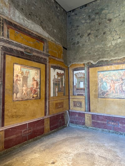 This image depicts an interior view of a room in Pompeii, Italy, showcasing well-preserved ancient Roman frescoes. The walls are adorned with vibrant paintings in yellow and red hues, featuring mythological scenes and architectural motifs.

On the left wall, a large fresco depicts a scene with several figures. A central male figure, seated and robed in red, appears to be gesturing. A smaller, nude figure, possibly a child, stands before him with snakes. Another figure, a woman, stands behind the seated man, while a fourth individual, dressed in a dark robe, is on the left, holding an object. Above this scene, architectural elements and decorative motifs are visible, including what appears to be an eagle sculpture.

To the right of this fresco, a narrow panel displays an illusionistic architectural rendering, creating a sense of depth with columns and a detailed interior structure. Further to the right, on the adjacent wall, another significant fresco shows a group of figures in dynamic poses, possibly depicting a mythological narrative involving several nude or semi-nude individuals, with one central male figure appearing to be a focal point of the action.

The lower portion of the walls is decorated with a deep red band featuring intricate patterns and smaller ornamental panels, including a depiction of mythological creatures in one such panel. The floor is composed of small, light-colored pebbles or tesserae, typical of Roman construction, and is partially illuminated by natural light entering from an unseen source. The upper sections of the walls transition to rougher, exposed stonework, suggesting the original construction before the plaster and frescoes were applied. The overall environment conveys a sense of ancient domestic or public space, offering a glimpse into the artistic and cultural practices of Pompeii.