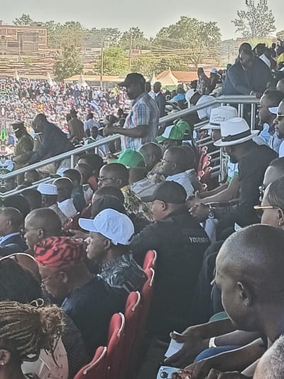This media file captures a large outdoor gathering of people, likely a public event such as a rally or meeting, set in Siaya, Kenya. Hundreds of individuals, predominantly adults and appearing mostly male, fill the frame, extending into the background.
In the foreground and mid-ground, people are standing and sitting on various structures, including what appear to be temporary metal barriers or bleachers. Many are casually dressed in shirts and t-shirts. Some individuals wear hats; one notable detail is a white fedora-style hat on which the text "FREEDOM OF SPEECH" is clearly visible. Another person in a black t-shirt has partially visible text that appears to read "YOURSELF". Red plastic chairs are visible in some sections, some occupied, others holding personal items.
The crowd is oriented towards a central, unseen point, suggesting a speaker or performance. In the background, beyond the immediate gathering, rows of people are visible, indicating the vastness of the crowd. Further back, there are trees, simple buildings, and a large white tent, possibly for shade or as a designated area. The overall lighting suggests it is daytime, likely late morning or afternoon, with clear weather conditions. The atmosphere appears attentive and engaged.