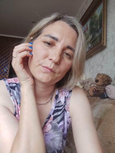 The image is a selfie of a woman with blonde hair, resting her hand on her cheek. She is wearing a floral-patterned top and a silver necklace. In the background, there is a framed painting on the wall, a teddy bear, and a beige couch. The room appears to be a living area. The presence of a painting, furniture, and domestic items suggests an indoor setting, possibly a home. There is no visual information in the image that definitively places the scene in Meleuz, Russia.
