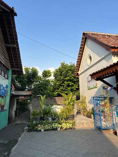 This is a medium shot of an outdoor scene in front of a building, likely a school, in Banguntapan, Indonesia, under a clear blue sky. The sunlight suggests it is late afternoon.

On the left, a building with a wooden facade and a tiled roof is visible, partially obscured by a wall with a mural. The mural is brightly colored, with images of clouds, letters, and numbers. A red and white Indonesian flag hangs from a pole attached to the building.

In the center, a walkway or path leads through an arched stone gateway, flanked by dense green trees. The path is paved with dark stones, and sunlight casts shadows on it.

On the right, a white building with a tiled roof and a decorative wooden eave is prominent. A circular window is visible high on its facade. A sign with text and images is mounted on the wall. Below the eave, a blue metal cage-like structure, filled with plastic bottles, stands on the paved ground. A collection of potted plants, including tall banana-like plants and smaller leafy plants, are arranged on a concrete step leading to the white building. Yellow vines hang from the eaves and one of the potted plants.

The ground in the foreground is paved with textured gray stones. The overall impression is one of a well-maintained, possibly educational, environment with a blend of natural elements and urban structures.