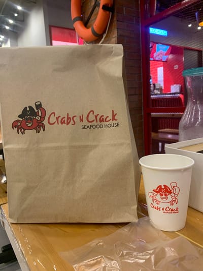 The image shows a close-up of a brown paper bag with the "Crabs N Crack Seafood House" logo printed on it in red. A cartoon crab wearing a pirate hat and eyepatch is also part of the logo. To the right of the bag is a white paper cup, also with the "Crabs N Crack" logo. The background shows a blurred interior scene with red and blue lighting, suggesting a restaurant or entertainment venue. There are also glimpses of what appear to be life preservers, possibly indicating a nautical theme. The overall impression is of a takeout order from a seafood restaurant in Angeles, Philippines. The lighting suggests it could be nighttime or an interior setting with artificial lighting.