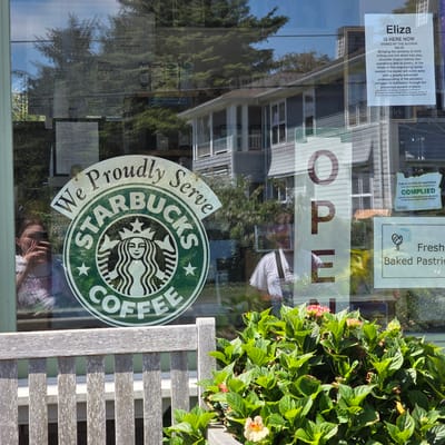 The image depicts a street-level view of a Starbucks store in Cannon Beach, Oregon, United States of America. The store's exterior window displays a large Starbucks logo with the text "We Proudly Serve Starbucks Coffee." The window reflects trees, buildings, and sky. A woman is visible through the window, taking a photo with her phone. To the right of the Starbucks logo, a large "OPEN" sign is displayed vertically. Further to the right, a sign indicates "Fresh Baked Pastries." Another sign, a "Public Notice of Sanitation Compliance," is also visible. In the foreground, a weathered wooden bench and lush green hydrangeas with pink and yellow flowers frame the view of the store. The overall scene suggests a sunny day in a coastal town.