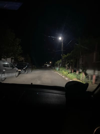 The image is a night-time, interior view from a vehicle driving through the city of Yasen, Bulgaria. The road is paved and appears to be a residential street. On the left side of the road, a silver car is parked, and further down, a black minivan is also parked. Trees and vegetation line the right side of the road, with some houses visible in the background. A street light illuminates the scene. The dashboard of the vehicle is visible in the foreground. The overall atmosphere is dark and quiet.