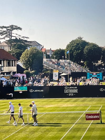 This media file depicts an outdoor tennis match, likely a professional tournament, as suggested by the banners and scoreboard. The scene is a manicured grass tennis court bathed in bright sunlight, indicating it is daytime with clear weather. A crowd of spectators is visible in tiered seating behind the court, some holding umbrellas for shade.
The main subjects are three male tennis players on the court, dressed in athletic attire and holding rackets. One player is mid-serve, while the other two appear to be positioned on their respective sides of the net, preparing to return the ball. The players are focused and engaged in the game.
Several objects are visible: the tennis net, the players' rackets, and a scoreboard displaying match scores. Prominent text on banners lining the court identifies the event. "LEXUS EASTBOURNE - OPEN -" is clearly displayed on a large black banner, along with the Lexus logo. Another banner on the net reads "Emirates". The scoreboard shows player names and scores, with a match seemingly in progress.
The backdrop includes lush green trees and the rooftops of buildings, suggesting the tennis court is located in an urban or suburban area with surrounding architecture. The overall atmosphere appears to be one of an active sporting event with spectators enjoying the competition under a sunny sky. The time of day is clearly daylight, given the strong shadows cast by the players and structures. The location context is specified as Brighton, United Kingdom.