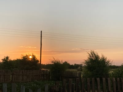 The image depicts a rural scene at sunset in Rakity, Russia. The sun is low on the horizon, casting a warm, orange glow across the sky and illuminating the clouds with soft, pastel hues. A rustic wooden fence runs horizontally across the foreground, with several wooden posts scattered in front of it. Behind the fence, lush green bushes and trees create a dense backdrop. A single wooden utility pole stands vertically on the right side of the image, with several horizontal wires stretching across the sky. The overall mood is serene and tranquil, evoking the peaceful atmosphere of a rural evening. No people are visible in the image.