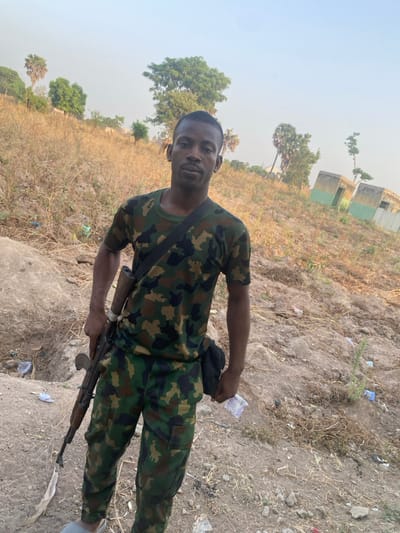 A man in camouflage fatigues poses outdoors, holding an AK-47-style rifle. He has a black bag slung across his body. The backdrop features a rural landscape with dry, grassy terrain, scattered trees, and distant buildings. The sky is hazy, suggesting possible dust or atmospheric conditions. The man appears to be in a field or clearing, with debris or litter visible on the ground.