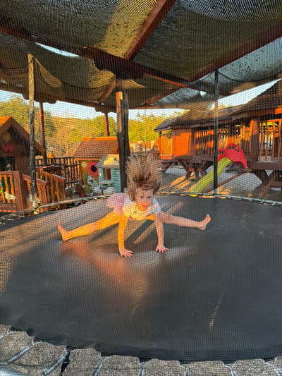 A young girl with blonde hair is captured mid-air, jumping on a trampoline. She is wearing a light-colored top and pink shorts, with her arms outstretched to her sides. Her hair is flying around her head, indicating the motion of her jump. The trampoline has a dark safety net surrounding it, and the springs connecting the mat to the frame are visible on the right side of the image.

Behind the trampoline, a multi-level wooden structure, possibly a playhouse or treehouse, is visible. This structure appears to be partially covered with a mesh netting, which casts shadows and adds texture to the scene. Various colorful objects, including what looks like a red toy and a yellow slide, are visible within the structure. The background suggests an outdoor setting with glimpses of trees and a hillside, bathed in the warm light of either sunrise or sunset. The overall mood is one of joyful activity and carefree play. The image was taken in Zagora, Montenegro.
