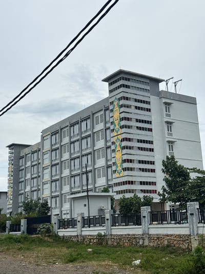 The image shows a multi-story building with a gray facade, situated in the city of Cotcut, Indonesia. The building has a modern architectural style with many windows. Decorative elements in yellow and green are present on one side of the building. The sky is overcast, suggesting a cloudy day. In the foreground, there is a fence and some overgrown vegetation. Power lines are visible in the upper portion of the image. The scene appears to be a street view of the building.