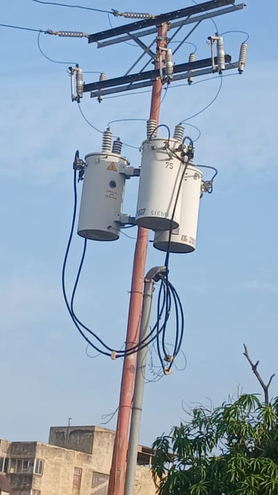 The image shows a power distribution system mounted on a pole, likely located in Barcelona, Venezuela, given the prompt's context. Three cylindrical electrical transformers, marked with "SIEMENS" and other identifiers, are horizontally mounted on a crossbeam attached to the pole. Insulators and electrical wires connect the transformers to the main grid running along the pole. Additional wires extend from the transformers towards buildings, including a multi-story concrete structure visible in the background. Some vegetation, possibly a tree, is present in the upper right corner of the frame. The sky is light blue.