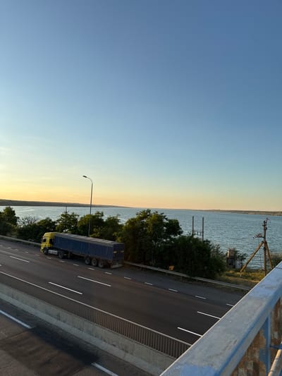 A yellow semi-trailer truck with a blue tarp-covered trailer is driving on a highway. The highway is lined with a concrete barrier and a metal fence. Beyond the highway is a body of water, possibly a river or lake, with trees on the opposite bank. The sky is clear and blue, with a hint of yellow from the setting sun. The scene appears to be in Yelyzavetivka, Ukraine, based on the provided context. There are no people visible in the image.