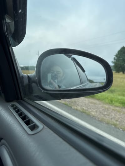 The image shows a view from inside a car, looking out the passenger window and into the side mirror. The side mirror reflects a person holding a phone to take a selfie. Outside the car, there is a rural landscape with a road and grassy fields. The sky is overcast. This scene is not located in the city of Arkhangel'skoe, Russia.