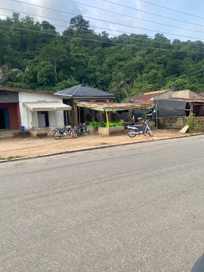 This image captures a street scene in the city of Igarra, Nigeria. In the foreground, a grey asphalt road occupies the lower portion of the frame, showing minor wear and light cracking. Adjacent to the road, a dirt shoulder or verge runs horizontally.

In the midground, a series of single-story buildings line the dirt verge. From left to right:
1.  A building with a grey and white painted facade, featuring a blue door on the left and a closed white panel or door in the center. A light-colored awning extends from the white section. A blue plastic barrel rests near its base.
2.  Three motorcycles are parked on the dirt in front of the buildings. Two darker-colored motorcycles (black/dark red) are positioned closer to the grey/white building. A lighter-colored motorcycle (white/blue/pink accents) is parked further right.
3.  Next to the first building, an open-sided structure features a dark, possibly corrugated, hip roof and a brightly colored, striped fabric awning (yellow, green, orange tones) covering an outdoor seating area. Several lime green plastic chairs and small tables are visible beneath this awning. Two indistinct human figures appear to be seated within this shaded area.
4.  Further to the right, a structure with a rusty corrugated metal roof and exposed concrete block walls is partially covered by a large black tarp or sheeting.
5.  Another structure, partially obscured, also with a rusty roof, is visible to the far right. A black plastic water tank is situated on the roof of one of the rightmost structures.

The background is dominated by a densely vegetated hill or small mountain, covered with various shades of green trees and foliage. Large boulders or rock formations are visible interspersed among the trees on the hillside, particularly on the left. The sky above is overcast with light grey clouds. Multiple thin, dark power lines stretch horizontally across the upper midground.