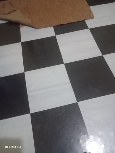 The image displays an indoor scene in Nkomo ward, Kenya. The prominent feature is a black and white checkered floor, likely composed of tiles or linoleum, exhibiting some surface wear and minor reflections. In the upper left portion of the frame, a brown, fibrous mat or rug is visible, partially uplifted from the floor, revealing its lighter-toned underside. No individuals, discernible actions, or interactions are present in the scene. A "REDMI 13C" watermark is visible in the lower-left corner of the image.