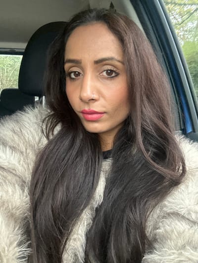A woman is depicted in a medium close-up portrait, seated inside a vehicle in Winslow, United Kingdom. She has medium-brown skin, dark brown eyes, and long, dark brown, wavy hair that extends over her shoulders. She is wearing a light gray or beige long-pile, fluffy garment. Her facial features are enhanced with eyeliner, mascara, light pink blush, and pink lipstick. A small silver nose ring is visible in her right nostril. She looks directly at the camera with a neutral expression. Behind her, a black car seat headrest is visible to the left, and a blue car door interior is partially visible to the right. Blurred green foliage, consistent with trees or bushes, is visible through the car window in the background. The scene is illuminated by natural daylight.