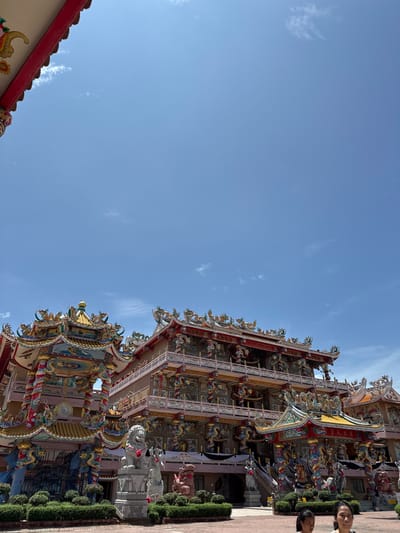In Ang Sila, Thailand, a vibrant, multi-tiered temple stands against a clear blue sky. The temple's architecture is highly ornate, featuring intricate dragon motifs, colorful sculptures, and tiered roofs adorned with gold and red trim. Two stone lion statues are positioned in front of the temple, one white and one reddish-brown, with red ribbons tied around their necks. Lush green bushes line the foreground, and a paved area in front of the temple is visible. In the lower right corner, two women are present, their faces partially visible. The temple appears to be a significant religious or cultural landmark.