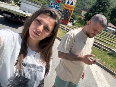 A bright, sunny upward-angled selfie captures a man and a woman at a roadside location, likely a gas station in Vetren Dol, Bulgaria. The image, taken in daylight with strong shadows, shows the subjects from the chest or waist up.
The man, positioned on the left, has close-cropped grey hair and a beard. He wears a light-colored t-shirt and green bottoms. Tattoos adorn his right forearm. He is holding what appears to be a hand-rolled cigarette or joint between his fingers, with his eyes closed in a relaxed expression.
The woman, on the right and closer to the camera, has long brown hair. She wears a white t-shirt with black lace detailing on the shoulder/collar, featuring the mirrored text "VERY INQUE" (likely "UNIQUE"). Her eyes are partially closed, possibly squinting in the bright sun, and she displays a subtle, relaxed smile.
The background reveals elements of a road and a gas station. A prominent yellow and red fuel price sign displays "МЕЧКАРОВ" (Mechkarov) at the top, with prices listed: "LPG 0.75", "DIESEL 1.71", "A95H 1.60", and "A100 1.60". Below the sign, a blue circular road sign with a white left-turn arrow, a red octagonal "STOP" sign, and a blue square sign with a fuel pump icon are visible. A black vehicle with the Bulgarian license plate "PA 1597 KP" is partially seen, along with parts of a yellow/green truck. The setting includes an asphalt road with a crosswalk, grassy verges, and a dense evergreen forest in the distance, consistent with the mountainous region of Vetren Dol, Bulgaria. The overall scene suggests a casual, relaxed stop during a journey.