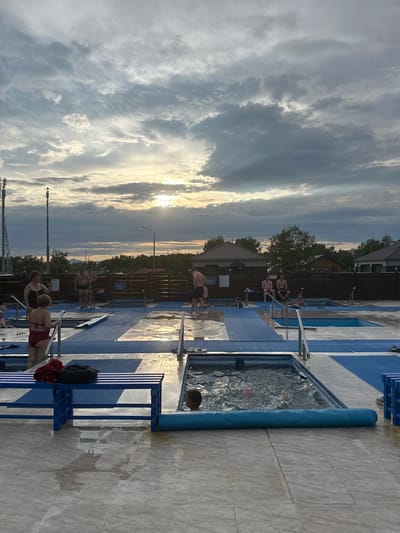 The image depicts an outdoor swimming pool area with several small, tiled pools. The pools are surrounded by blue matting and light-colored tiled flooring, which is wet in places, indicating recent use. Several people are present, some are in the pools, while others are standing or sitting around them. The sky is cloudy with the sun visible through the clouds, casting a warm light. In the background, there are buildings, trees, and telecommunication towers, suggesting an urban or semi-urban setting, possibly in Yuzhnyye Koryaki, Russia.