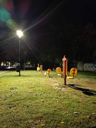The scene depicts a public park area at night in Tolosa, Argentina. A tall street light on the left side provides the primary illumination, casting bright white light across the green grassy foreground and creating prominent lens flare and long shadows. Adjacent to the street light, a dark-colored cylindrical waste bin is mounted on a pole. Diagonally extending from the mid-ground towards the upper right, a series of at least three distinct pieces of outdoor exercise equipment are visible. These units feature red/orange vertical support structures and yellow plastic seats or footrests. The ground consists of green grass with scattered patches of bare earth and fallen yellow-brown leaves. In the background on the right, dense dark foliage partially obscures a wall with visible markings or graffiti. On the far left, in the distant background, several additional streetlights and parked vehicles are discernible along what appears to be a paved road. No individuals or active interactions are present in the depicted environment.
