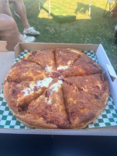 A large pizza, cut into eight slices, sits in a cardboard box lined with green and white checkered paper. The pizza has a thick, golden-brown crust and appears to be topped with cheese and some type of meat. The box is open, and the pizza is the primary subject in the foreground.
In the background, out of focus, are people's legs and feet. One person is wearing white Crocs, and another is wearing patterned Crocs. They are sitting on a grassy area, likely outdoors. To the left of the people, a portion of a cornhole board can be seen on the grass. In the further background, there are shadows cast by what appear to be outdoor chairs and possibly a tent or umbrella. The overall scene suggests a casual outdoor gathering. The context of "city of AB, Canada" is not visually present in the image.